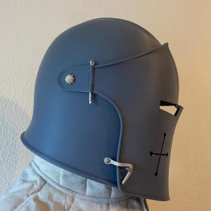 May include: A steel blue helmet with a visor and a cross design on the front. The helmet has a rounded top and a hinged visor. The visor has a small opening for the eyes. The helmet is designed for protection.