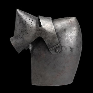 Pode incluir: Uma se&ccedil;&atilde;o de armadura de metal antiga, provavelmente um componente de capacete, contra um fundo preto. A pe&ccedil;a tem uma forma curva com uma viseira perfurada e uma al&ccedil;a. O metal tem uma apar&ecirc;ncia desgastada e envelhecida.