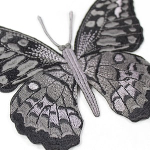 Op de afbeelding: Geborduurde vlinderpatch in grijstinten en zwart. De vlinder heeft gedetailleerde vleugelpatronen en een getextureerde uitstraling. De antennes en het lichaam zijn lichtgrijs, wat een contrast vormt met de donkere vleugelranden.