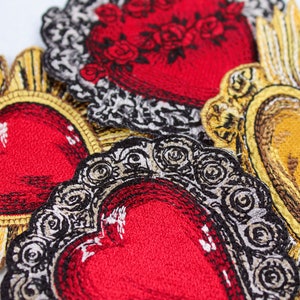 Puede incluir: Primer plano de parches bordados con corazones rojos con marcos ornamentados en oro y negro. Los corazones est&aacute;n superpuestos, creando un dise&ntilde;o visualmente atractivo e intrincado.