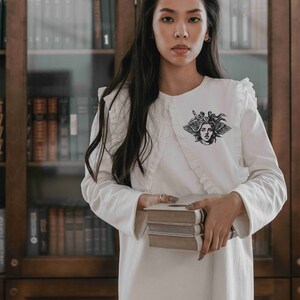May include: A white long-sleeved dress with a ruffled collar and a black Medusa graphic. The dress is held by a person holding three books. The background features a wooden bookshelf filled with books.