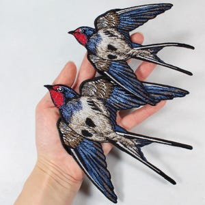 May include: Two embroidered swallow patches. The birds have blue wings, brown and white bodies, and red heads. The patches are detailed with black outlines and long tail feathers. The patches are likely for sewing onto clothing or accessories.