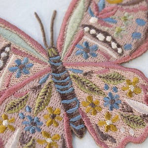 Op de afbeelding: Een geborduurde vlinderpatch in tinten roze, bruin, blauw, groen en geel. De vlinder heeft bloemmotieven en is perfect om een vleugje whimsy toe te voegen aan knutselwerk en projecten.