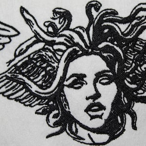 May include: White textile featuring a black embroidered design of Medusa's face with snakes for hair and winged elements. The detailed embroidery creates a striking contrast against the white background, showcasing the mythical figure.