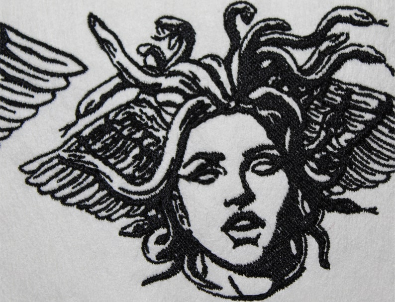 May include: White textile featuring a black embroidered design of Medusa's face with snakes for hair and winged elements. The detailed embroidery creates a striking contrast against the white background, showcasing the mythical figure.
