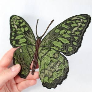 Op de afbeelding: Geborduurde vlinderpatch in groen- en zwarttinten. De vlinder heeft gedetailleerde vleugelpatronen en een bruin lichaam en antennes. De patch is waarschijnlijk bedoeld voor decoratieve doeleinden, zoals op kleding of accessoires.
