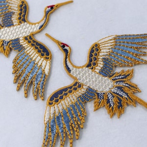 May include: Two embroidered iron-on patches of blue and gold cranes with white accents. The cranes are in flight with their wings spread.