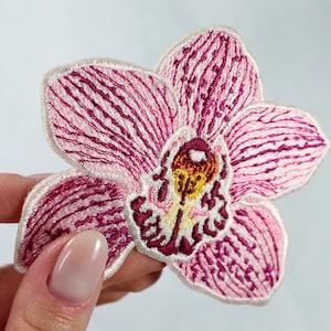 May include: Embroidered orchid flower patch with pink and white petals and a yellow and maroon center. The patch has detailed stitching and a white border. This floral patch can be used to decorate clothing or accessories.