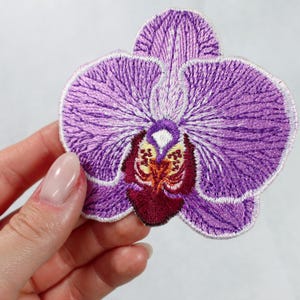 May include: Embroidered purple orchid flower patch. The patch features detailed stitching in shades of purple, with a contrasting center in burgundy, yellow, and white. The flower's petals are fully visible, showcasing the intricate design.