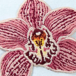 May include: Embroidered orchid patch in shades of pink and burgundy. The flower has detailed stitching, with a cream-colored border. The center of the flower features yellow and orange accents. The petals have a textured appearance.