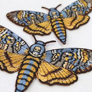 Peut inclure: Écussons brodés représentant un papillon tête de mort. Les écussons présentent des détails complexes avec une palette de couleurs bleue, jaune, marron et grise. Les ailes du papillon sont déployées et le design est très détaillé.