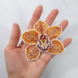May include: Embroidered orchid patch in shades of orange, yellow, and brown. The flower has detailed stitching and a white border. This floral patch can be used to decorate clothing, bags, or other fabric items.