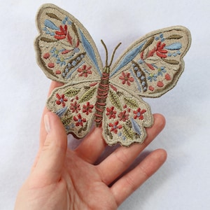 Op de afbeelding: Een beige geborduurde vlinderpatch met rode, blauwe en groene bloemdetails. De vlinder is ongeveer 10 cm breed en 7,5 cm hoog.