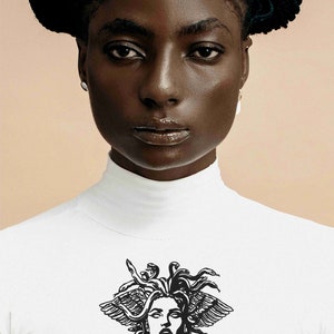 May include: A white turtleneck shirt featuring a black graphic of Medusa's head. The shirt is worn by a person with dark skin and hair. The background is a solid, light tan color.