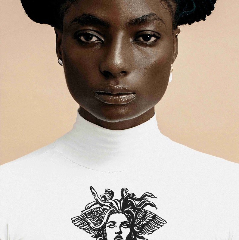 May include: A white turtleneck shirt featuring a black graphic of Medusa's head. The shirt is worn by a person with dark skin and hair. The background is a solid, light tan color.