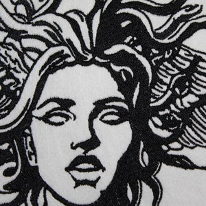 May include: Close-up of a white textile with a black embroidered design of a woman's face with snakes for hair. The design is highly detailed, with intricate lines and shading, creating a striking contrast.