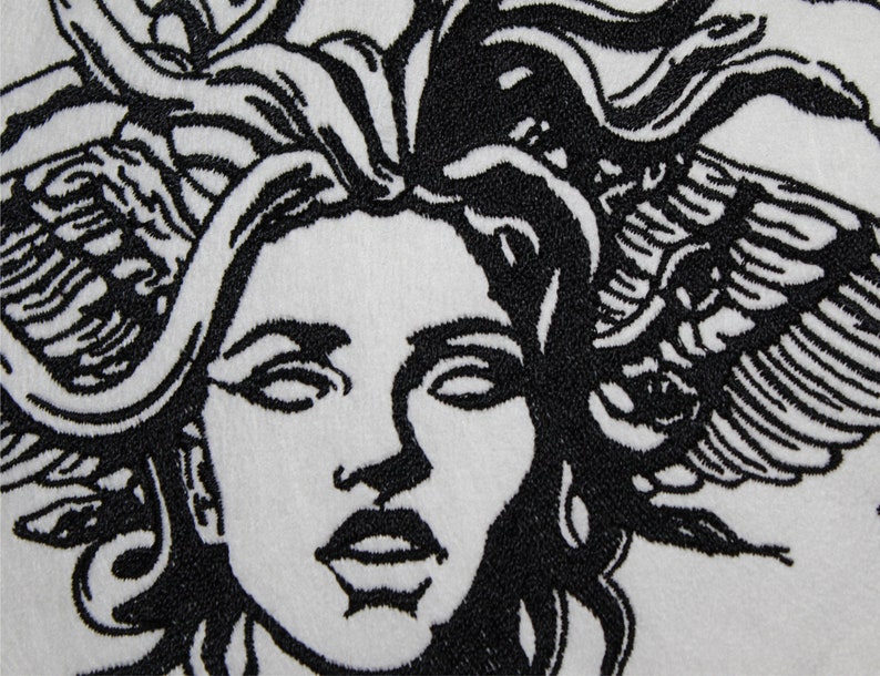May include: Close-up of a white textile with a black embroidered design of a woman's face with snakes for hair. The design is highly detailed, with intricate lines and shading, creating a striking contrast.