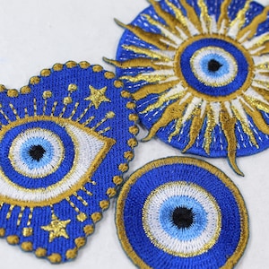 May include: Embroidered patches featuring the evil eye symbol in blue, white, and gold. One patch is heart-shaped, another is circular, and the third is a sunburst design. The patches are detailed with gold thread and star accents.