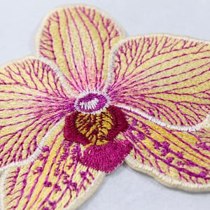Może przedstawiać: Zbliżenie haftowanej naszywki w kształcie kwiatu orchidei. Płatki są w kolorze miękkiej żółci z delikatnymi fioletowymi żyłkami. Środek kwiatu jest w kolorze głębokiej magenty, ze skomplikowanymi detalami ściegów. Naszywka jest prawdopodobnie przeznaczona do odzieży lub projektów rzemieślniczych.
