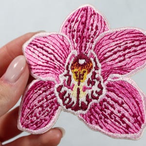 Puede incluir: Parche bordado de orquídea en tonos rosa, blanco y amarillo. La flor presenta costuras detalladas y un aspecto texturizado. El parche está diseñado para coser o planchar sobre tela, agregando un elemento floral decorativo.