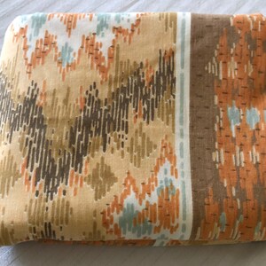 May include: Folded fabric with a repeating geometric pattern in shades of tan, brown, orange, and light blue. The design features vertical and zigzag lines, creating a textured appearance. A brown border runs along one edge.