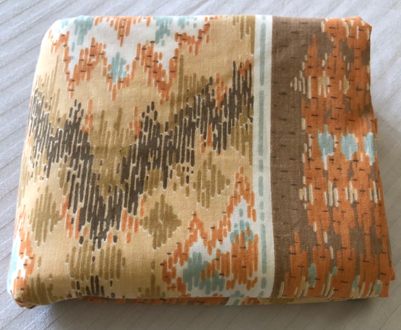 May include: Folded fabric with a repeating geometric pattern in shades of tan, brown, orange, and light blue. The design features vertical and zigzag lines, creating a textured appearance. A brown border runs along one edge.