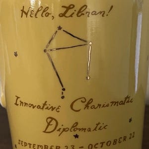 May include: A yellow ceramic mug with the text "Hello, Libran!" and the Libra constellation in dark brown. Additional text includes "Innovative, Charismatic, Diplomatic" and "September 23 - October 22".