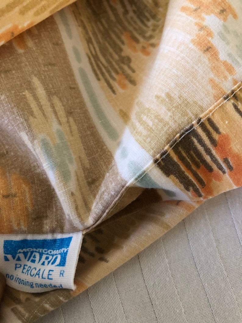 May include: Close-up of a patterned fabric with a tag. The fabric features a design in shades of brown, tan, orange, and blue. The tag reads "MONTGOMERY WARD PERCALE R no ironing needed."
