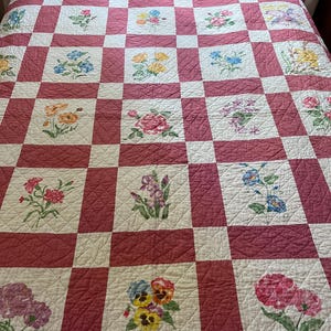 May include: A handmade quilt with a red and white checkered pattern. Each white square features a hand-painted floral design in various colors. The quilt is likely made of cotton and is suitable for a full or queen-sized bed.