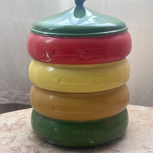 May include: A stacked, colorful container with a lid. The container is composed of four sections: green lid, red, yellow, and olive-green sections. The lid has a small, dark blue handle. The container sits on a marble-patterned surface.