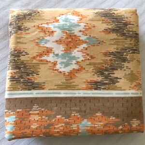May include: Folded fabric with a geometric pattern in shades of tan, brown, orange, and teal. The design features diamond and linear shapes. A brown border with orange accents is visible at the bottom.