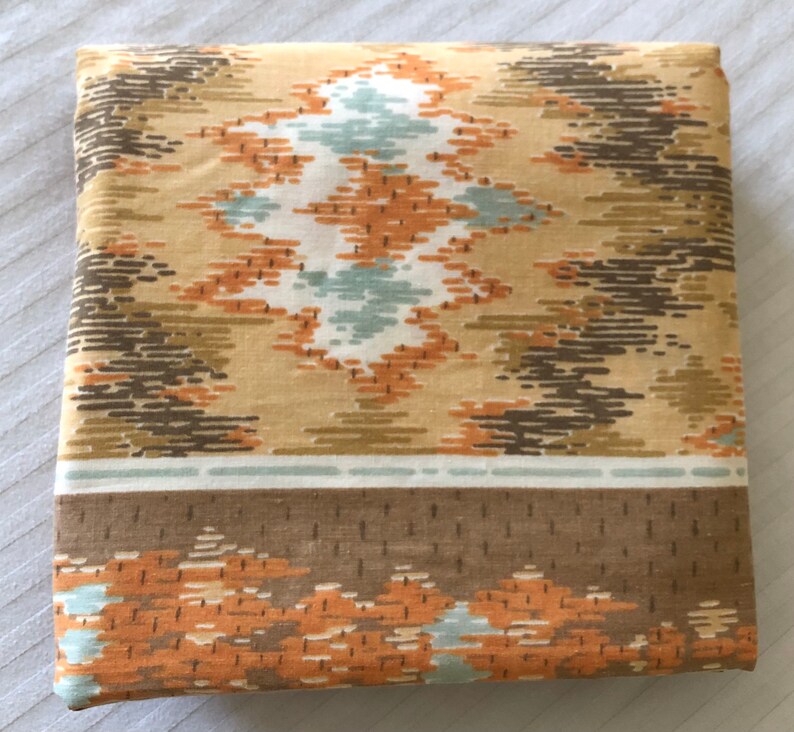 May include: Folded fabric with a geometric pattern in shades of tan, brown, orange, and teal. The design features diamond and linear shapes. A brown border with orange accents is visible at the bottom.