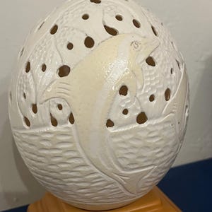 May include: A white, carved eggshell sculpture featuring a dolphin design. The eggshell has a textured surface with cut-out holes and is mounted on a brown wooden base. The dolphin is carved in relief.