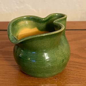 May include: A small, green ceramic pitcher with a unique, wavy design. The pitcher has a light yellow glaze on the inside.
