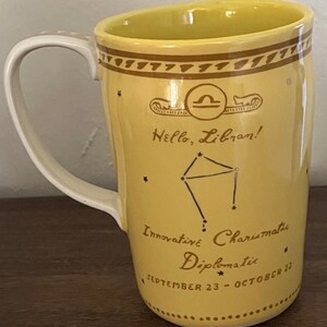 May include: Yellow ceramic mug with a white handle and interior. The mug features a Libra zodiac design with the text "Hello, Libran!" and the dates "September 23 - October 22."