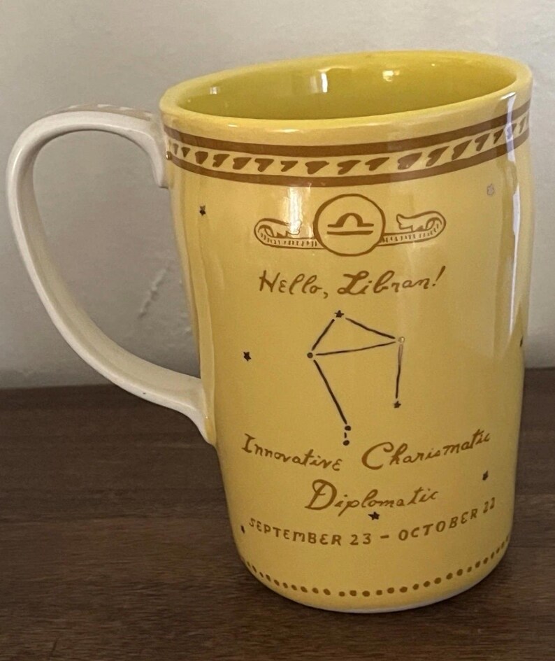 May include: Yellow ceramic mug with a white handle and interior. The mug features a Libra zodiac design with the text "Hello, Libran!" and the dates "September 23 - October 22."