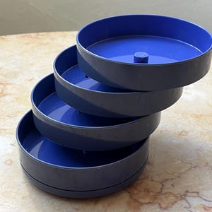 May include: A stack of four blue and gray plastic trays. Each tray has a dark gray exterior and a bright blue interior. The trays are round and appear to be designed to nest together. The background is a light marble surface.