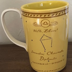 May include: Yellow ceramic mug with a white handle. The mug features a Libra zodiac symbol, constellation, and the text "Hello, Libran!" along with the traits "Innovative, Charismatic, Diplomatic" and the dates "September 23 - October 22."