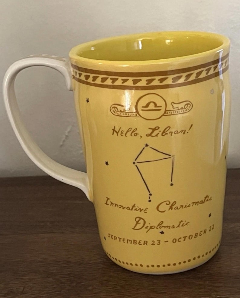May include: Yellow ceramic mug with a white handle. The mug features a Libra zodiac symbol, constellation, and the text "Hello, Libran!" along with the traits "Innovative, Charismatic, Diplomatic" and the dates "September 23 - October 22."