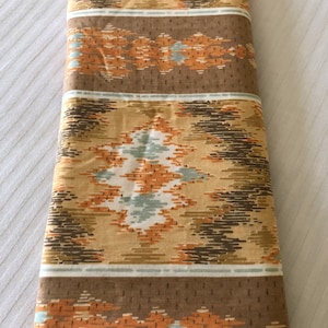 May include: Folded fabric with a geometric pattern in shades of tan, brown, orange, and teal. The design features diamond shapes and horizontal stripes, creating a vintage or retro aesthetic. The fabric appears to be cotton or a similar material.