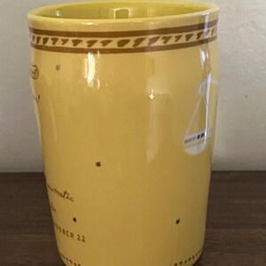 May include: Yellow ceramic mug with a brown patterned band at the top and a Libra zodiac symbol. The mug has text that reads "climatic" and "OCTOBER 22" with star accents. The mug is on a wooden surface.