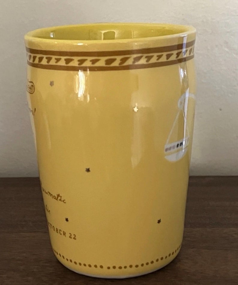 May include: Yellow ceramic mug with a brown patterned band at the top and a Libra zodiac symbol. The mug has text that reads "climatic" and "OCTOBER 22" with star accents. The mug is on a wooden surface.