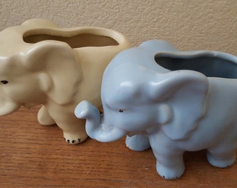 Puede incluir: Dos macetas de cerámica con forma de elefante, una en esmalte azul claro y la otra en esmalte crema. Ambas tienen un agujero en la parte superior para plantar.