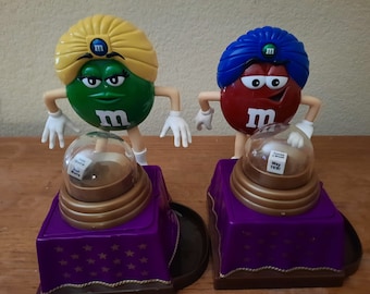 以下が含まれることがあります： 2人のM&Mキャラクターが占い師に扮しています。緑のM&Mは黄色のターバンをかぶり、青と白のサイコロが入ったガラスの球を持っています。赤いM&Mは青いターバンをかぶり、中に「Way to go!」と書かれた白い紙が入ったガラスの球を持っています。両方のキャラクターは、金色の縁取りと茶色の土台が付いた紫色のプラットフォームの上に立っています。