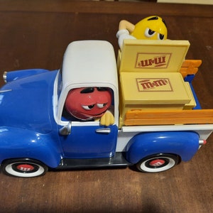Puede incluir: Un camión de juguete azul y blanco con personajes de M&M's. Un personaje M&M's rojo está en el asiento del conductor, y un personaje M&M's amarillo está en la caja del camión. La caja del camión contiene cajas amarillas con el logotipo de M&M's.