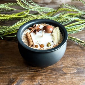 May include: A black ceramic candle holder filled with a white candle. The candle is decorated with a cinnamon stick, star anise, a bay leaf, and other spices. Greenery is in the background.