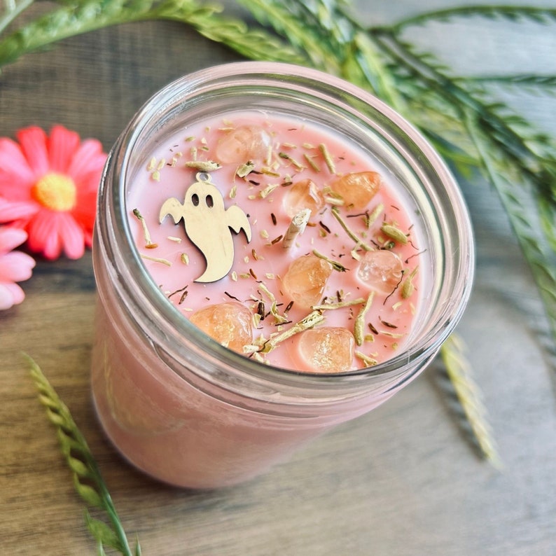 May include: A pink candle in a clear glass jar topped with dried herbs, several translucent orange crystals, and a small silver-toned ghost charm. The candle features a single centered wick and is filled with smooth, light pink wax.