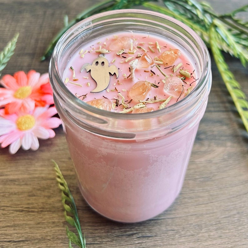 May include: A pink candle in a clear glass jar topped with small orange crystals, dried herbs, and a silver-colored metal ghost charm. The wax surface is smooth and light pink, featuring a single centered wick surrounded by decorative elements.