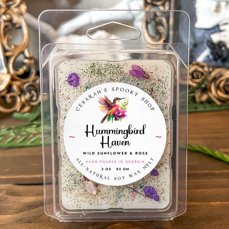 May include: A wax melt in a clear plastic container. The wax is off-white with green glitter and purple flower accents. The label reads "Hummingbird Haven" with a hummingbird illustration. It is 3 oz (85 grams) and made with all-natural soy wax.