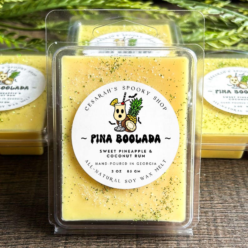 May include: Yellow wax melt bars in clear plastic packaging. The label reads "Pina Boolada" with a cocktail and pineapple graphic. The wax melt is scented with sweet pineapple and coconut rum, hand-poured in Georgia, and made with all-natural soy wax.